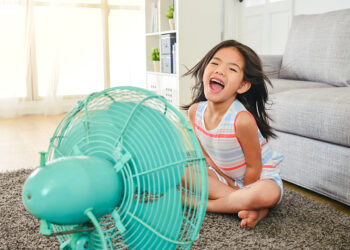 litle cute girl laughing and sitting in front of fan. refreshing herself from hot weather with legs crossed.