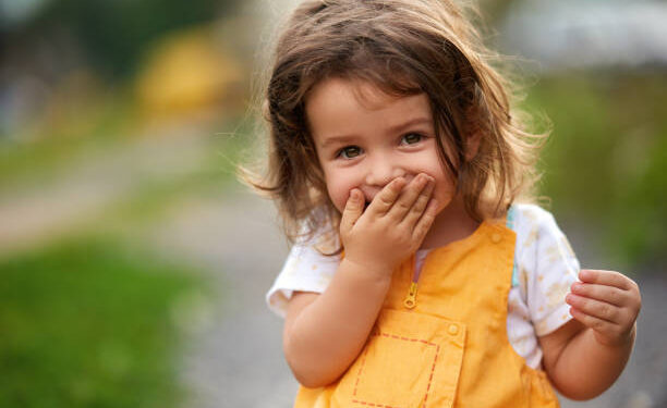 Little girl with brown hair outdoors, covering her mouth laughing