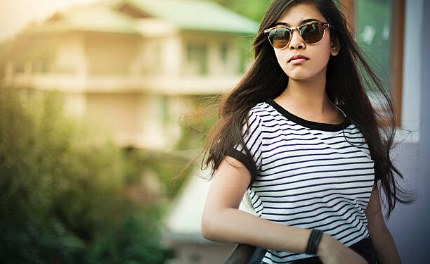 Outdoor image of beautiful, happy late teen urban girl enjoying fresh air in balcony at day time. She is wearing sunglasses, her long hair are flying in air. One person, horizontal composition with selective focus and copy space.